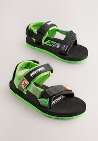 Juodos ir žalios spalvos sandalai su reguliuojamomis dirželėmis ir tekstūruotais padais. Ant jų yra „Minecraft“ prekės ženklo ženklai ir ryškiomis spalvomis pavaizduoti personažai.