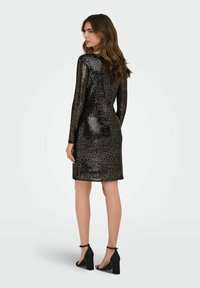 Robe noire à sequins avec une silhouette ajustée, manches longues, longueur mi-cuisse et décolleté rond. Associée à des escarpins noirs.