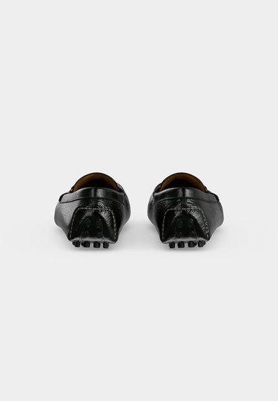 Paire de mocassins de conduite en cuir noir montrée par le talon avec des picots en caoutchouc sur la semelle et à l’arrière pour l’adhérence.