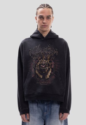 Jonge persoon met gevlochten haar draagt een zwarte hoodie met een gedetailleerde afbeelding van een hand die een brandend hart vasthoudt en vervaagde blauwe spijkerbroek.