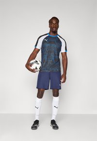 Zwart en blauw gestreept sportshirt, navy korte broek, witte sokken, zwarte voetbalschoenen; holding een voetbal, op een neutrale achtergrond.
