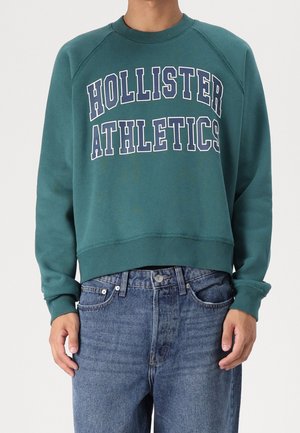 Osoba ubrana w turkusową bluzę Hollister Athletics i niebieskie dżinsy, stojąca na jednolitym jasnym tle.