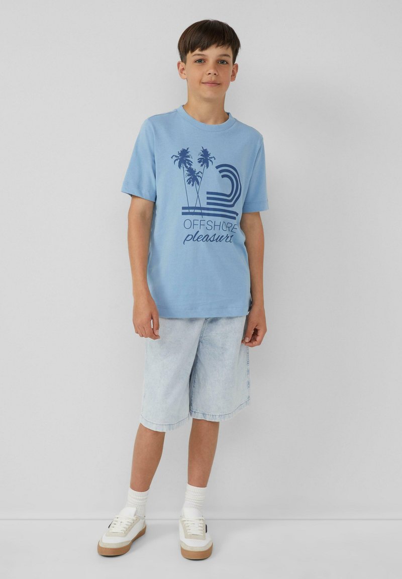 Hellblaue Baumwoll-T-Shirt mit grafischem Design von Palmen und Text; kombiniert mit hellen Jeans-Shorts und weißen Turnschuhen mit beigen Akzenten.