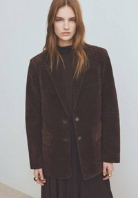 Blazer in corduroy marrone con revers a punta, due tasche frontali e due bottoni. Indossato sopra un dolcevita nero, con una consistenza morbida.