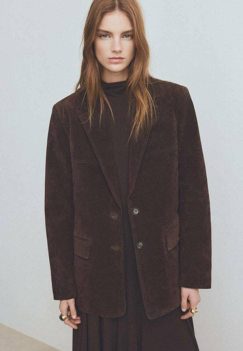 Blazer in corduroy marrone con revers a punta, due tasche frontali e due bottoni. Indossato sopra un dolcevita nero, con una consistenza morbida.