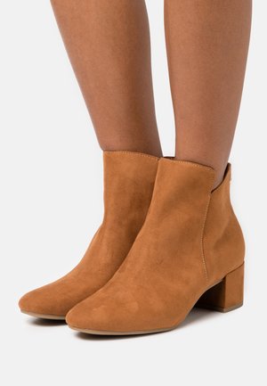 Femme portant des bottines en suède beige mi-mollet avec de petits talons bloc, texture lisse et orteils ronds sur fond blanc.