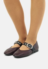 Scarpe piatte bordeaux con un cinturino nero caratterizzato da accessori in argento e dettagli con occhielli. Texture liscia con punta arrotondata e tacco basso.