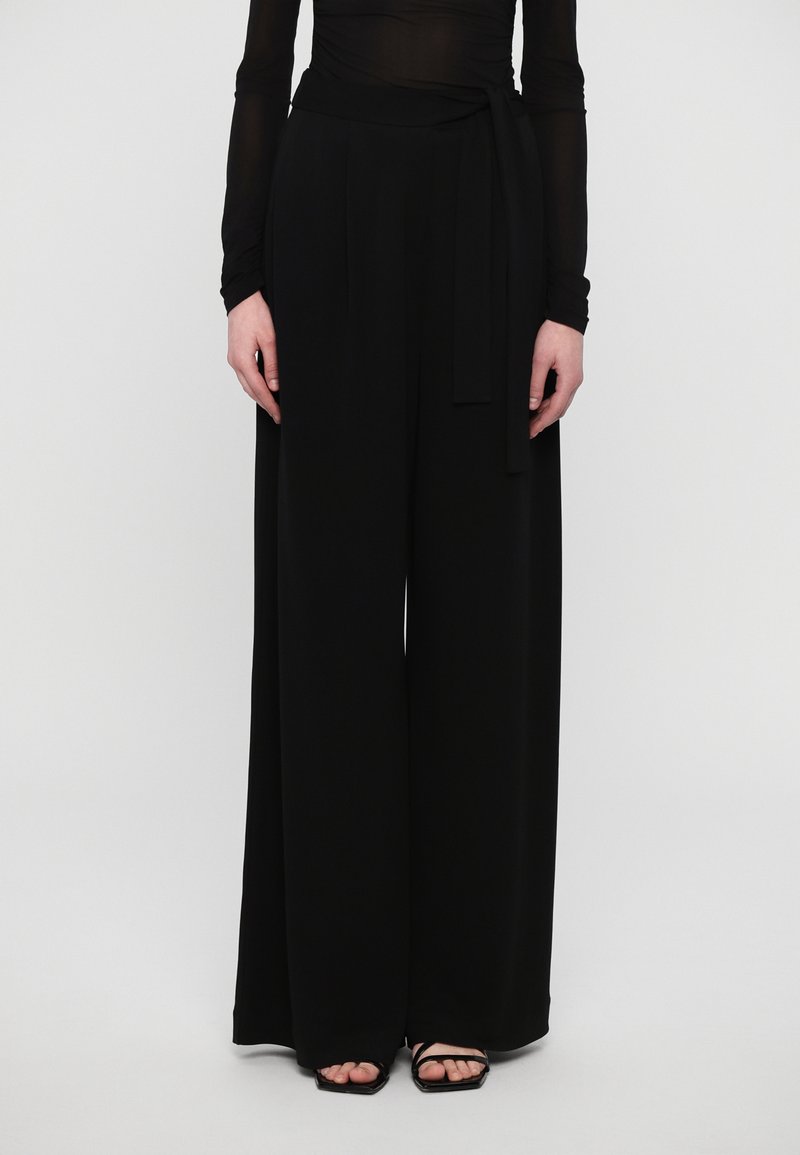 Wide-leg black trousers with a soft texture, featuring a high waist and a tied belt detail. Paired with black open-toe heels.