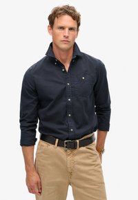 Marineblauw overhemd met knopen, een borstzak, opgerolde mouwen en beige broek. Het overhemd heeft een zachte textuur en subtiele knopen.