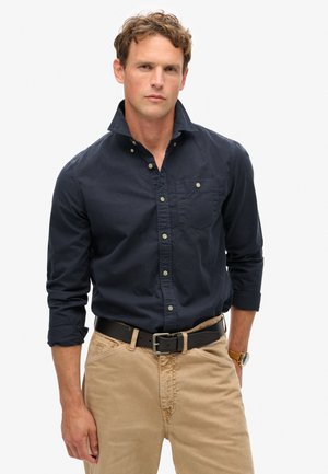 Marineblauw overhemd met knopen, een borstzak, opgerolde mouwen en beige broek. Het overhemd heeft een zachte textuur en subtiele knopen.