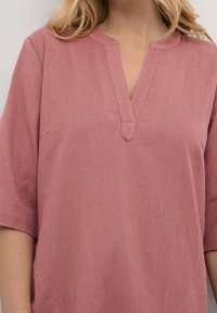 Rosa Leinentunika mit V-Ausschnitt und kurzen Ärmeln. Sie hat eine dezente Textur und eine leicht A-linienförmige Silhouette. Keine sichtbaren Muster oder Verzierungen.