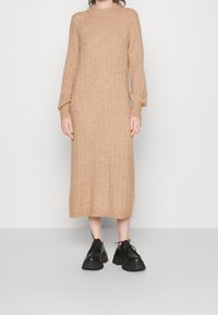 Robe pull en tricot beige avec texture côtelée, manches longues et col montant. Chaussures noires à lacets épaisses. Fond neutre.
