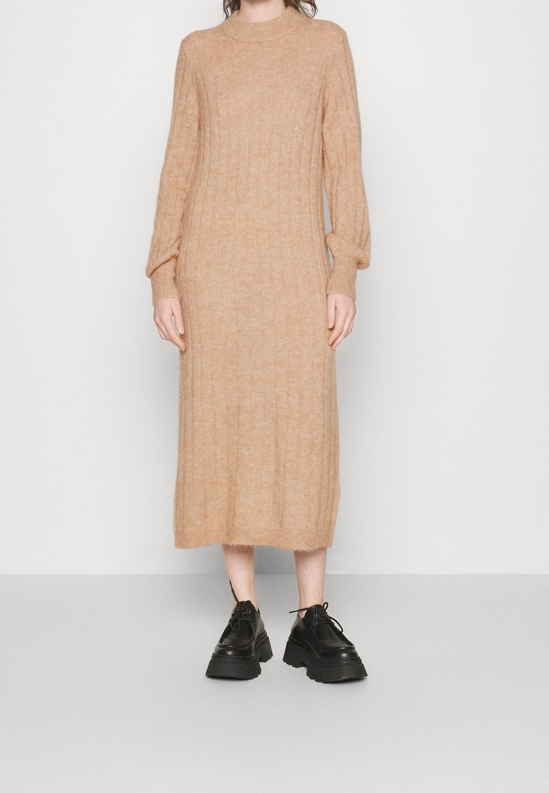 Robe pull en tricot beige avec texture côtelée, manches longues et col montant. Chaussures noires à lacets épaisses. Fond neutre.