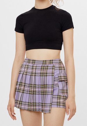Kobieta w czarnym krótkim topie z odkrytym brzuchem i liliowej plisowanej spódnicy w kratę z boczną kieszenią i ozdobnym pierścieniem.