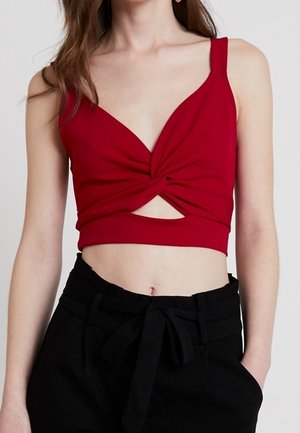 Frau trägt ein rotes, ärmelloses Crop-Top mit verdrehtem Frontdetail und schwarze, hoch taillierte Hose mit gebundenem Gürtel.
