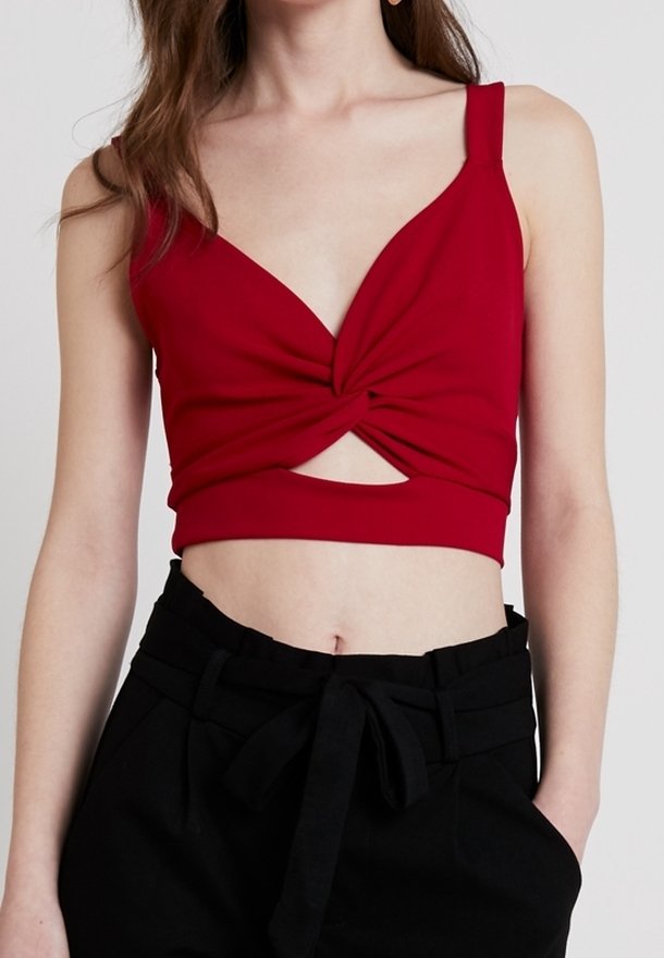 Femme portant un crop top rouge sans manches avec un détail torsadé à l'avant et un pantalon noir taille haute avec une ceinture nouée.