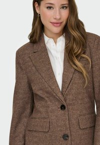 Bruine tweed blazer met een geprofileerd patroon, met notch revers, twee voorzakken en twee zwarte knopen. Witte blouse met kraag eronder.