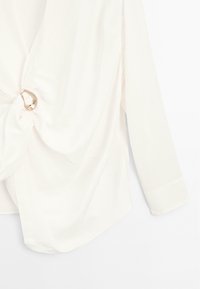 Blouse en soie blanche avec un détail de nœud torsadé et un accent en anneau argenté. Elle possède des manches longues et une texture lisse et fluide.