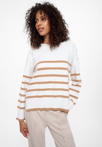 Pullover bianco con strisce orizzontali color beige, orlo a coste e bottoni sulle spalle, abbinato a pantaloni beige chiaro. Capelli ricci visibili.