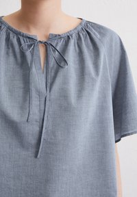 Top azzurro chiaro a maniche corte realizzato in tessuto morbido. Presenta un collo arricciato con un'apertura a chiave e un dettaglio a fiocco.