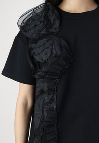 Černé bavlněné tričko s průsvitným černým volánem na levé straně. Volán má texturovaný, vrstvený vzhled. Krátké rukávy.