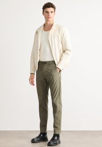 Jonge man in een beige jack, wit geribbeld tanktop, olijfgroene broek met trekkoord en zwarte instappers, staand tegen een effen achtergrond.