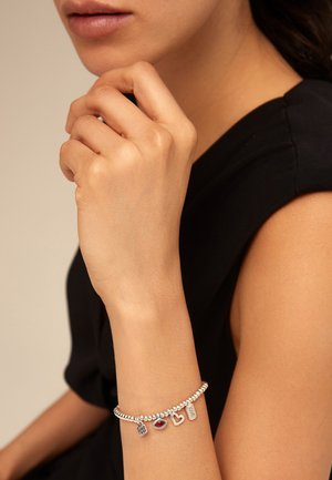 Pulsera de plata con cuentas esféricas y charms, que incluyen un labio rojo, un corazón y una etiqueta grabada con la palabra "amor", mostrada en una mano.
