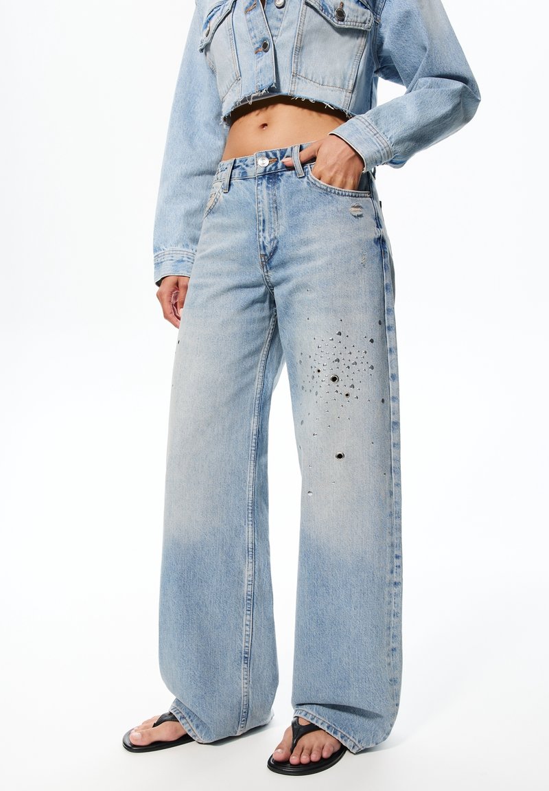 Personne portant un jean bleu clair à jambes larges avec des trous usés et des ornements, une veste en denim courte et des tongs noires.