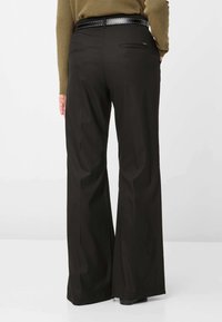 Zwarte broek met wijde pijpen gemaakt van gladde stof; heeft twee achterzakken en een riem met metalen studs, gestyled met een groene trui.