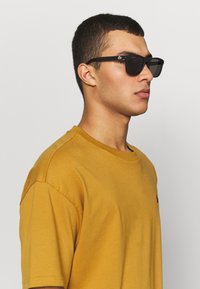 Ochelari de soare negri cu rame pătrate și lentile întunecate, purtați de o persoană îmbrăcată cu un tricou galben muștar, evidențiind un design simplu, dar modern.