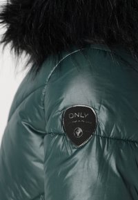 Green puffer jacket featuring a shiny, smooth finish, black faux fur trim on the hood, and a textured black logo patch on the sleeve.