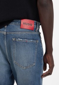 Blugi din denim albastru închis cu o curea de piele roșie etichetată „HUGO” în talie, cu buzunare standard și detalii uzate.