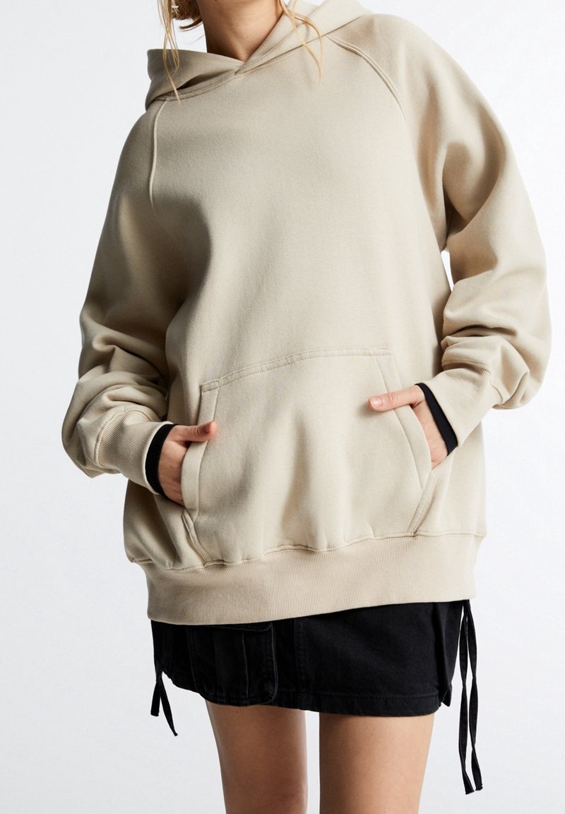 Personne portant un sweat à capuche beige oversize avec poche avant et jupe noire, mains glissées dans les poches du sweat.