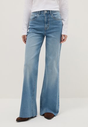 Vrouw draagt lichtblauwe high-waist wijde jeans met ruwe zomen en bruine puntige schoenen, gecombineerd met een wit topje met lange mouwen.