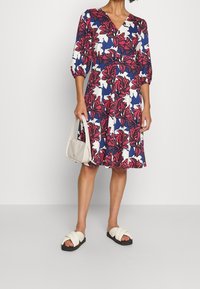 Vestido con estampado floral que presenta un escote en V, mangas de tres cuartos y una falda acampanada. Los colores incluyen azul marino, blanco y burdeos. Combinado con un bolso crema.