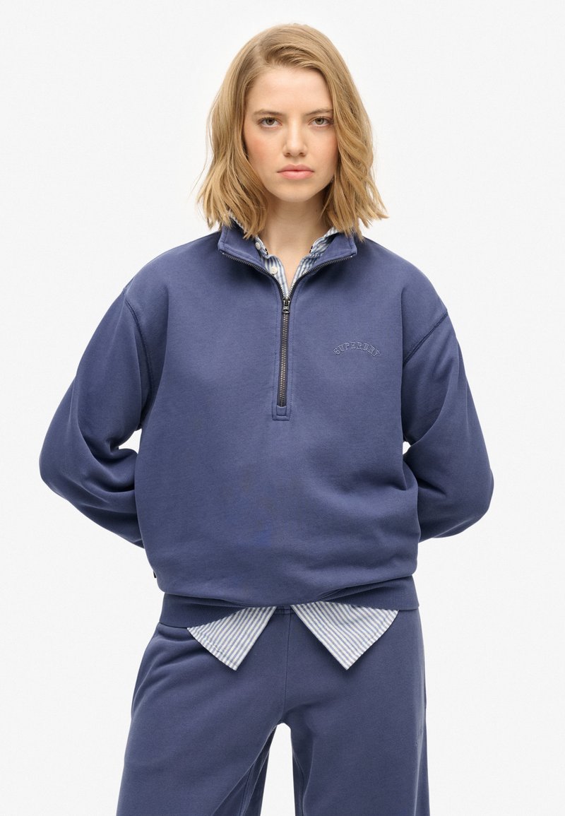 Granatowy polarowy pulower z półsupełkiem i haftowanym logo, z obniżonymi ramionami i prążkowanym wykończeniem. Nałożony na paskowaną koszulę z kołnierzykiem.