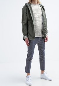 Olivgrön, huva-jacka med tryckknappar, lager ovanpå en strukturerad ljusgrå tröja. Grå slim-fit byxor och vita sneakers.