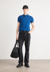 Kék polo póló fehér kiemelésekkel, fekete farmer, fekete sportcipő és fekete táska. A modell egy világos betonpadlón áll, semleges háttérrel.
