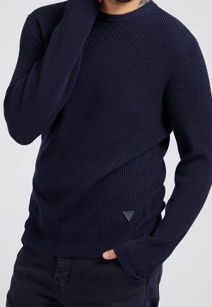 Man die een donkerblauwe, getextureerde gebreide trui draagt met lange mouwen en geribbelde manchetten, gecombineerd met een donkere broek, hand in de zak.