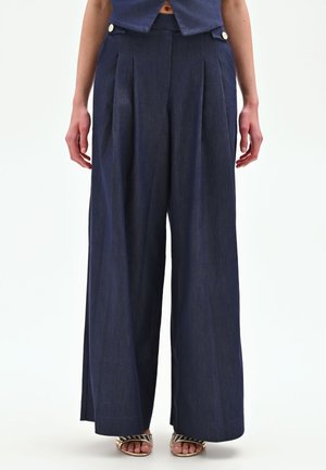 Donna che indossa pantaloni blu navy a vita alta e gamba larga con pieghe e dettagli con bottoni, abbinati a sandali metallici open toe.