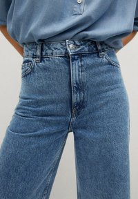 Jean en denim bleu taille haute avec une texture lisse, design à cinq poches, et des boutons argentés à l'avant.