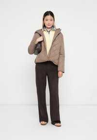 Jachetă maro matlasată cu glugă, bluză tricotată galbenă și pantaloni maro cu un croi larg. Modelul ține o geantă mică maro. Fundal deschis.