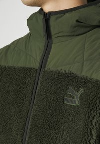 Veste zippée verte avec un dessus matelassé, un bas en polaire douce et un logo Puma noir sur la poitrine. Le tissu a une texture moelleuse.