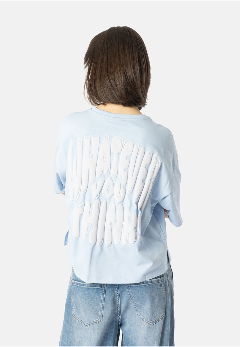 Jasnoniebieska, oversize koszulka z białą teksturowaną grafiką na plecach z napisem "COKOLWIEK MYŚLISZ!" w zaokrąglonym kroju.