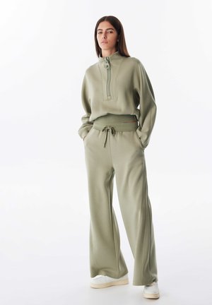 Felpa verde chiaro con zip e colletto alto, abbinata a pantaloni a gamba larga coordinati. Tessuto morbido, vestibilità casual e vita con cordoncino.