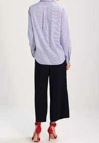 Modrá a bílá pruhovaná košile s dlouhými rukávy, detaily na knoflících a rozparkem vzadu, v kombinaci s širokými černými kalhotami a červenými botami na podpatku.