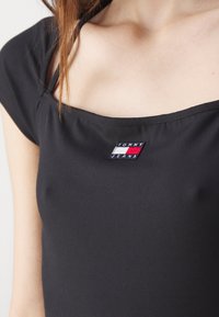 Svart ribbad topp med kvadratisk halsringning, med en liten broderad "TOMMY JEANS"-etikett i röd, vit och blå på bröstet.