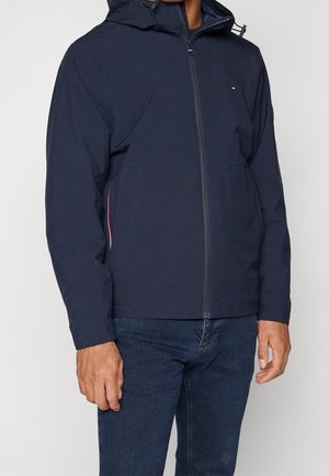 Homme portant une veste à capuche bleu marine zippée avec un petit logo sur la poitrine et un jean bleu foncé, debout devant un fond uni.