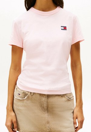 Femme portant un t-shirt rose pâle à manches courtes avec un petit logo Tommy Jeans et une jupe en jean beige avec des détails de poches.