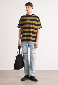 Gestreepte korte mouw shirt in mosterd en zwart met een zon grafiek, gecombineerd met versleten lichtblauwe jeans en zwarte schoenen, met een zwarte tote bag.